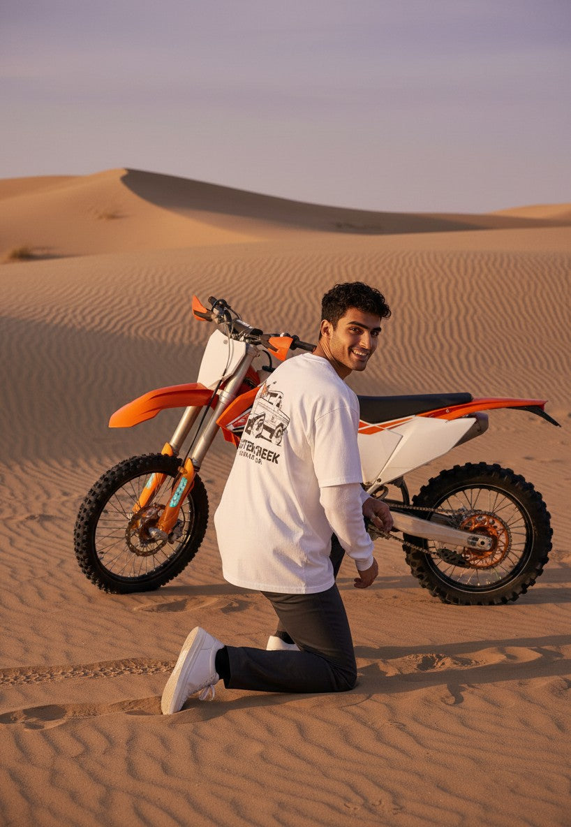 Man wearing a white Scatter Creek Trading Co long sleeve shirt kneeling in the desert next to an orange dirt bike